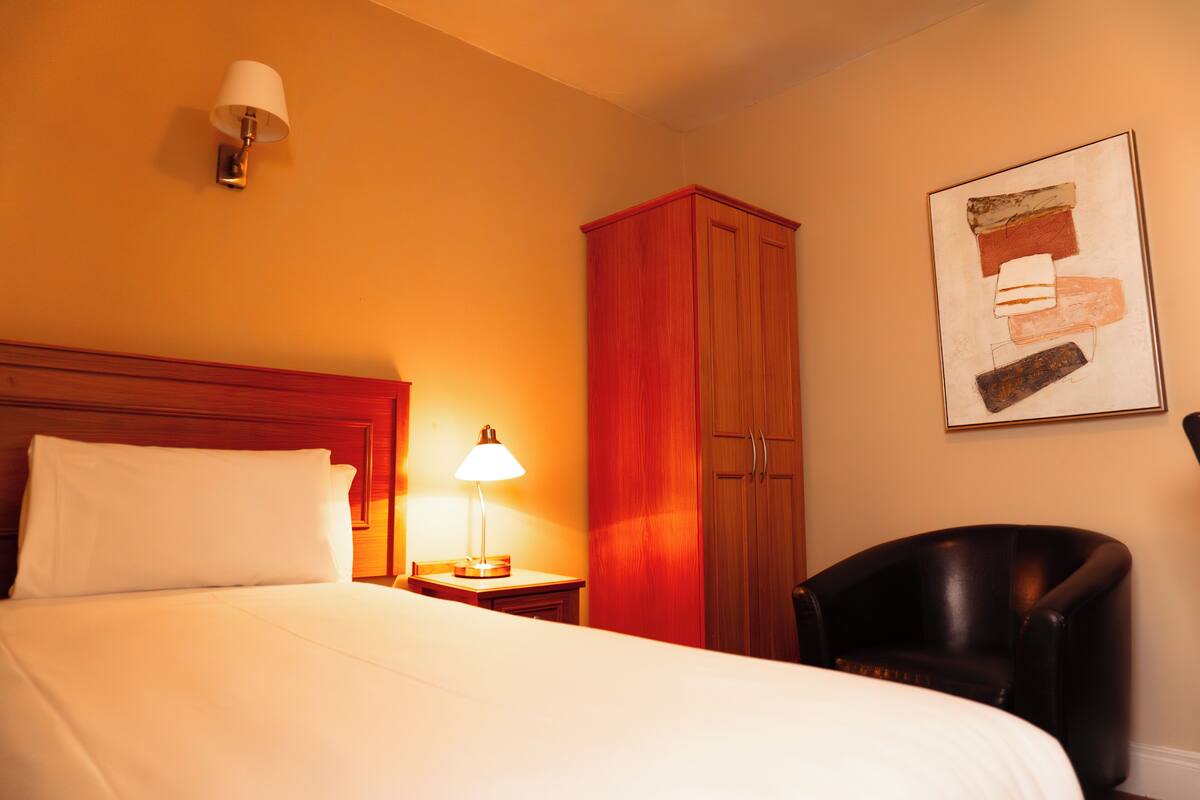 A hotel room with a bed, a nightstand with a lamp, a wooden wardrobe, a framed artwork on the wall, and a leather armchair.