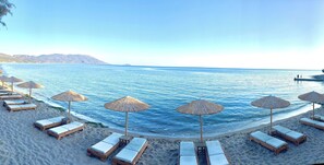 A beach with wooden lounge chairs and thatched umbrellas, a calm sea, and distant mountains. - Votsalakia Hotel (Samos)