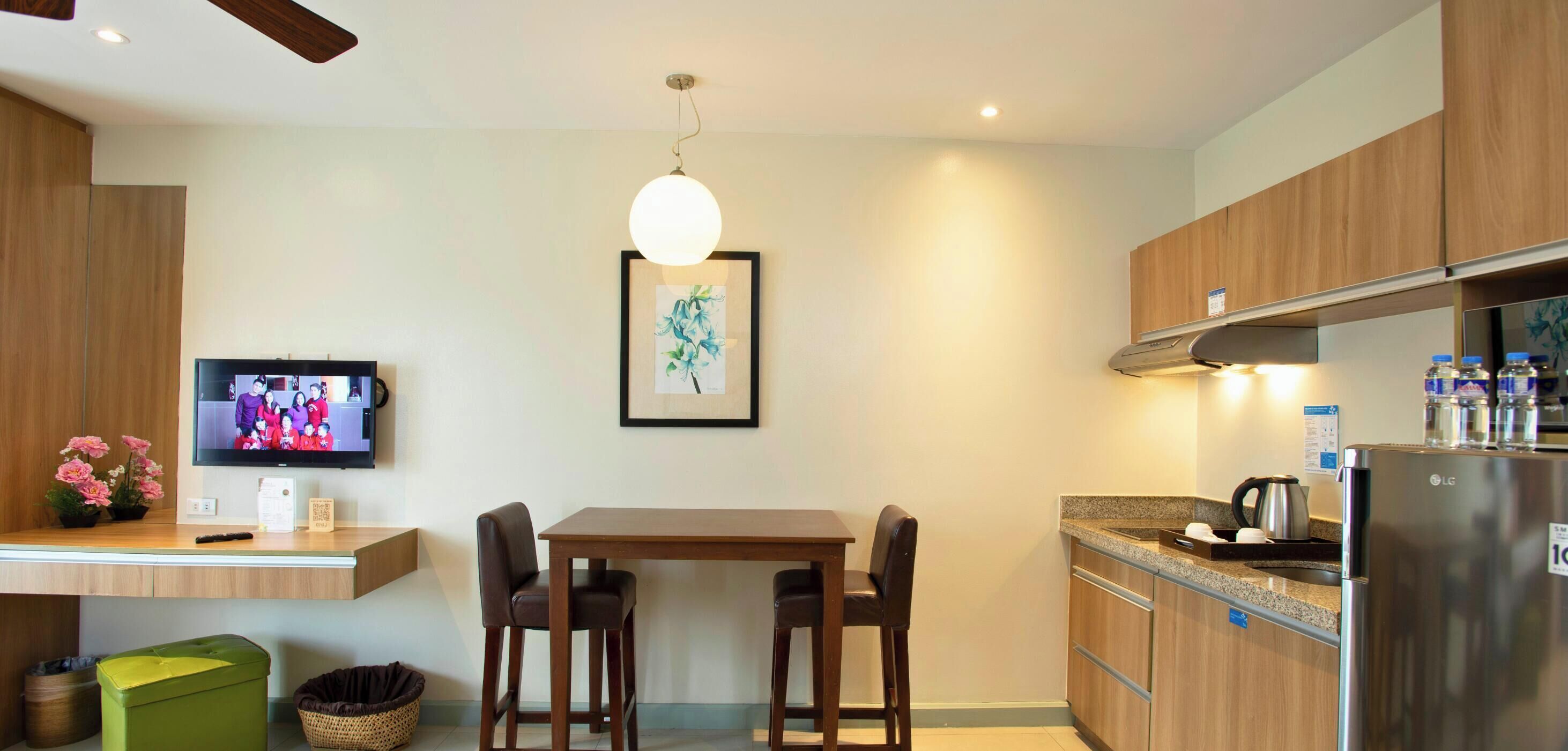 Una cocina moderna con armarios de madera, un refrigerador de acero inoxidable, una mesa de comedor con sillas, un televisor en la pared y una lámpara colgante.