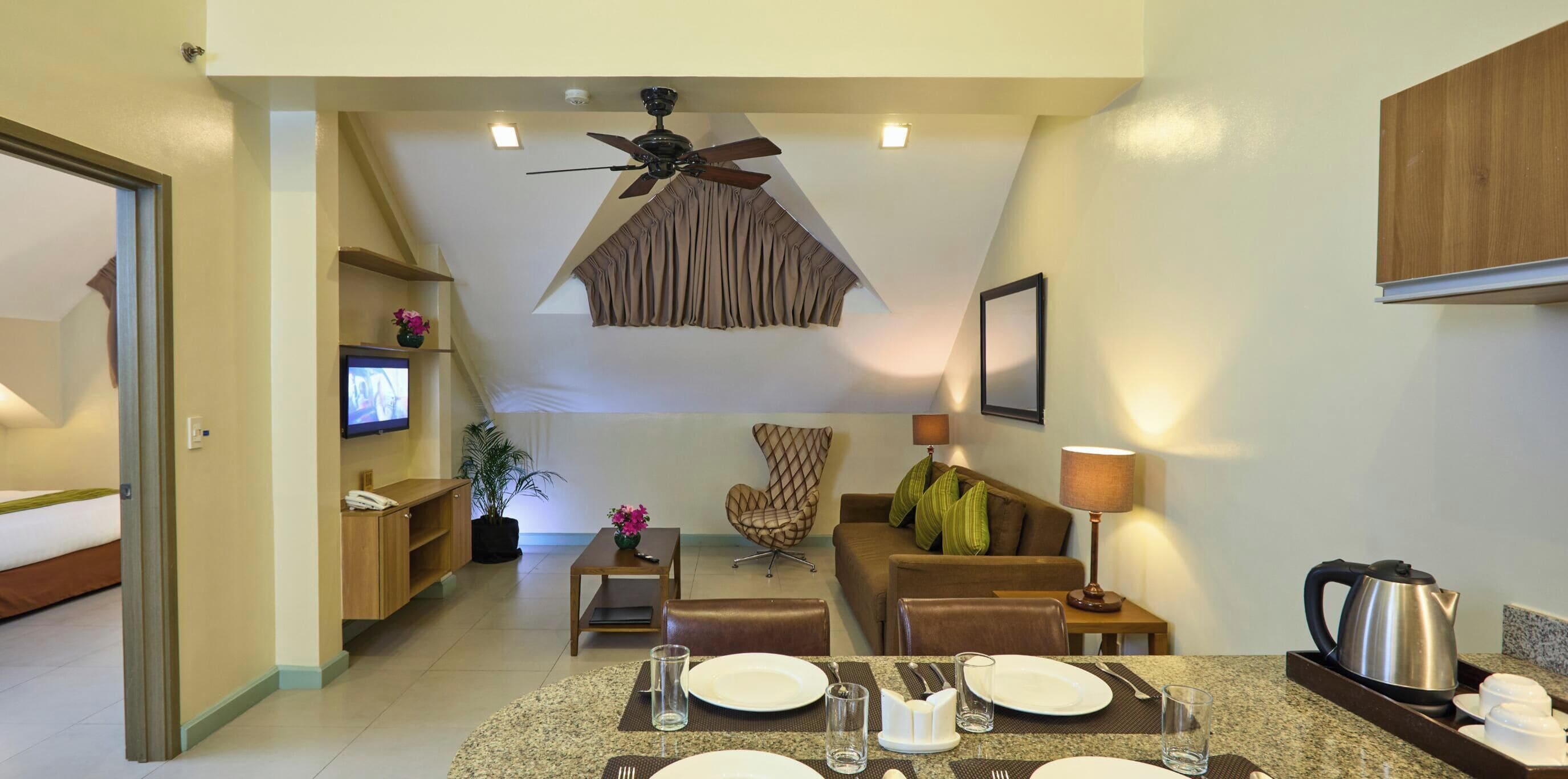 A hotel room with a dining area, a sofa, a TV, a ceiling fan, and a kitchenette.