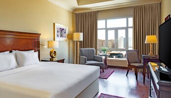 Una habitación de hotel con una cama grande, un escritorio con televisor, una silla, una mesita con una planta y un ventanal con cortinas.
