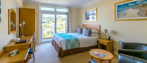 A hotel room with a bed, a desk, a chair, a small table, and a balcony with a view.