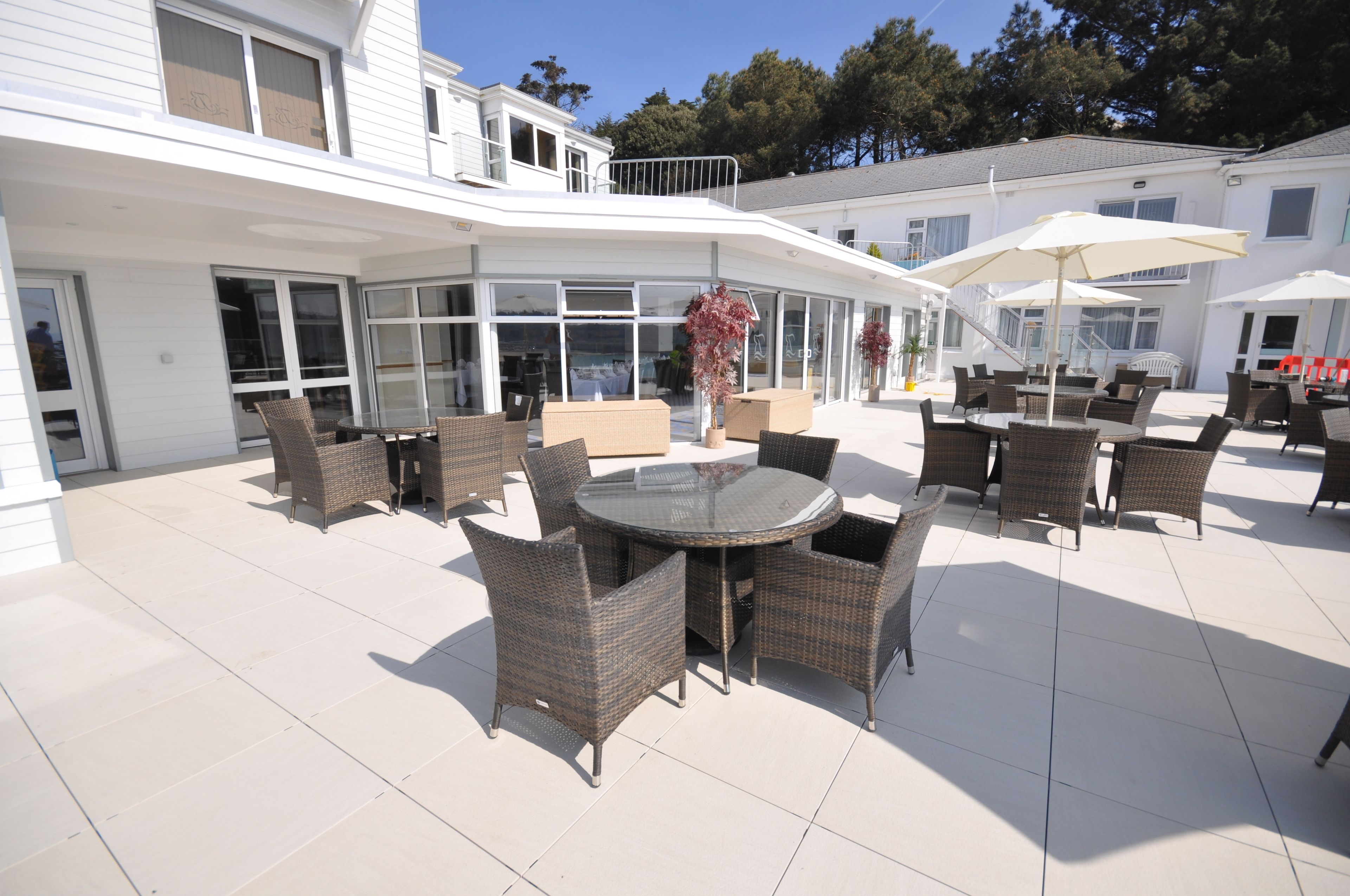 Un patio techado al aire libre con mobiliario de mimbre y sombrillas, ventanas amplias y un edificio blanco al fondo.