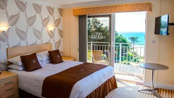A hotel room with a bed, a nightstand, a TV, and a balcony with a view of the sea.
