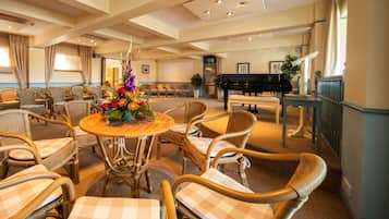 Un amplio hall de hotel con sillones y mesas de mimbre, un piano de cola y una gran composición floral.