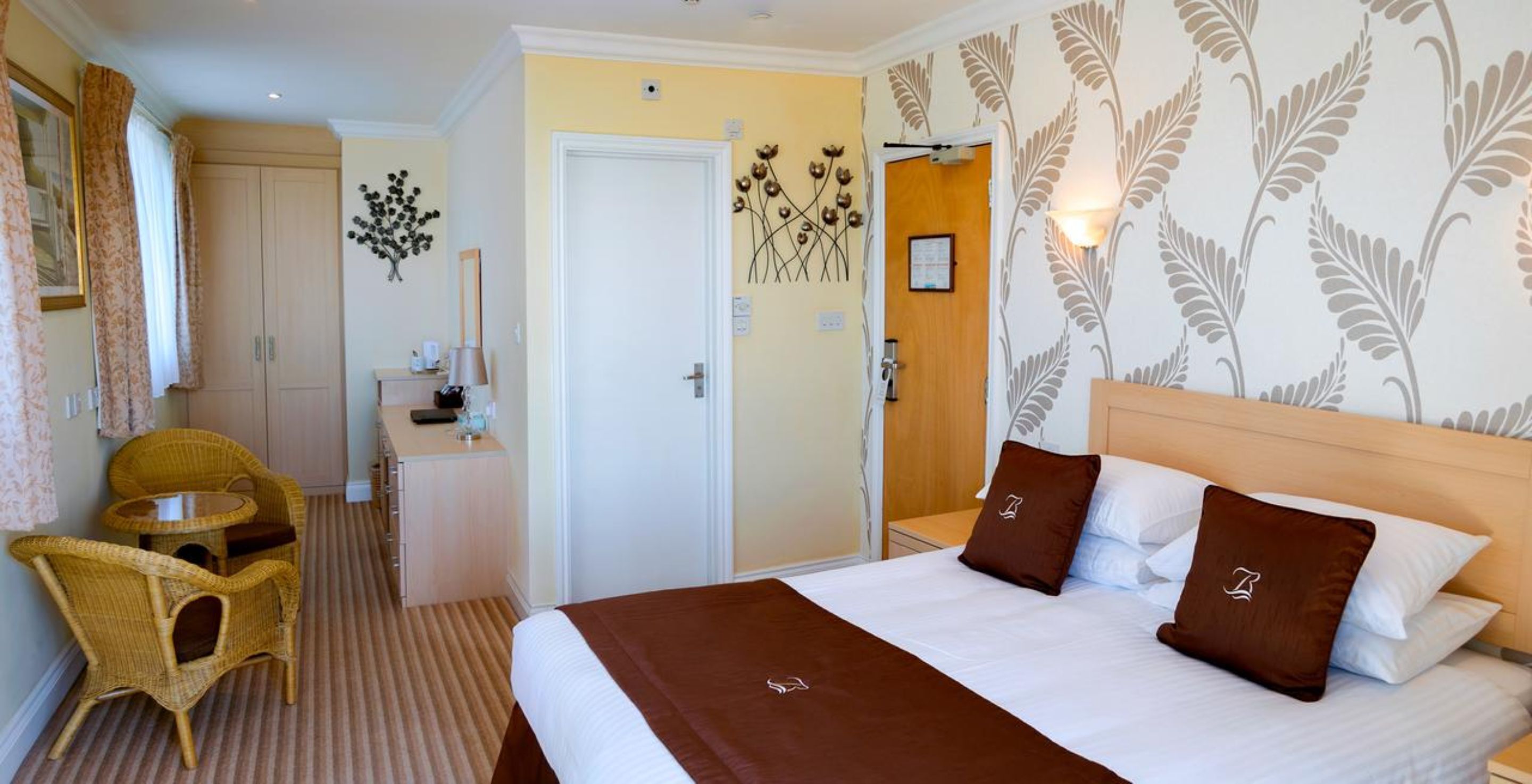 A hotel room with a bed, two brown pillows, a wooden headboard, a bedside table, a chair, a desk, and a door leading to another room.