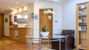 Un área de recepción con una mesa de vidrio, una orquídea en maceta, un sillón de cuero marrón, una estantería de madera y un mostrador de recepción con encimera de madera.