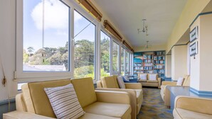 Una habitación con sofás color beige, una estantería y ventanas amplias con vistas a árboles.