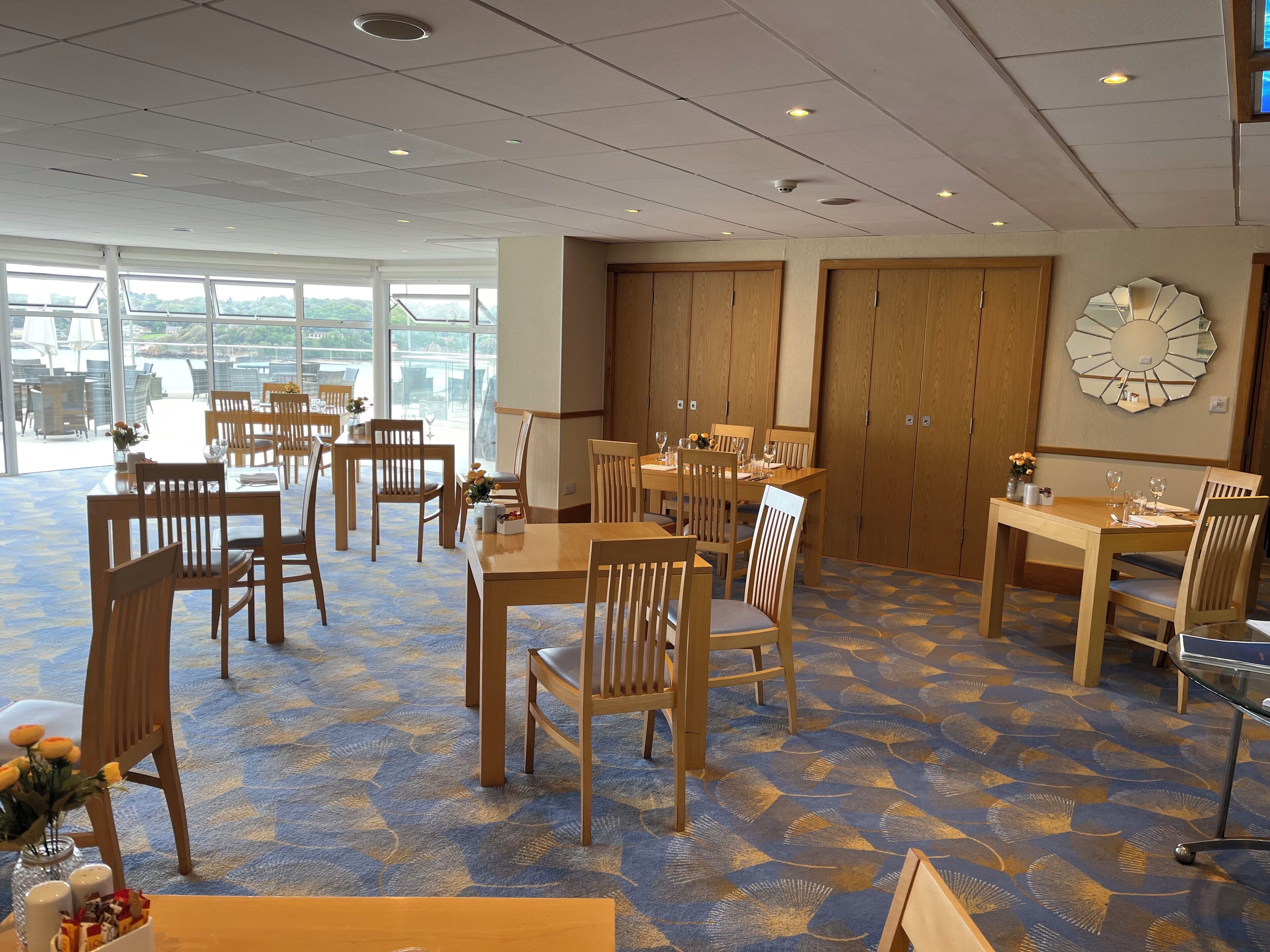 A spacious dining area with wooden tables and chairs, large windows offering a view of the outdoors, and a ceiling with recessed lighting.