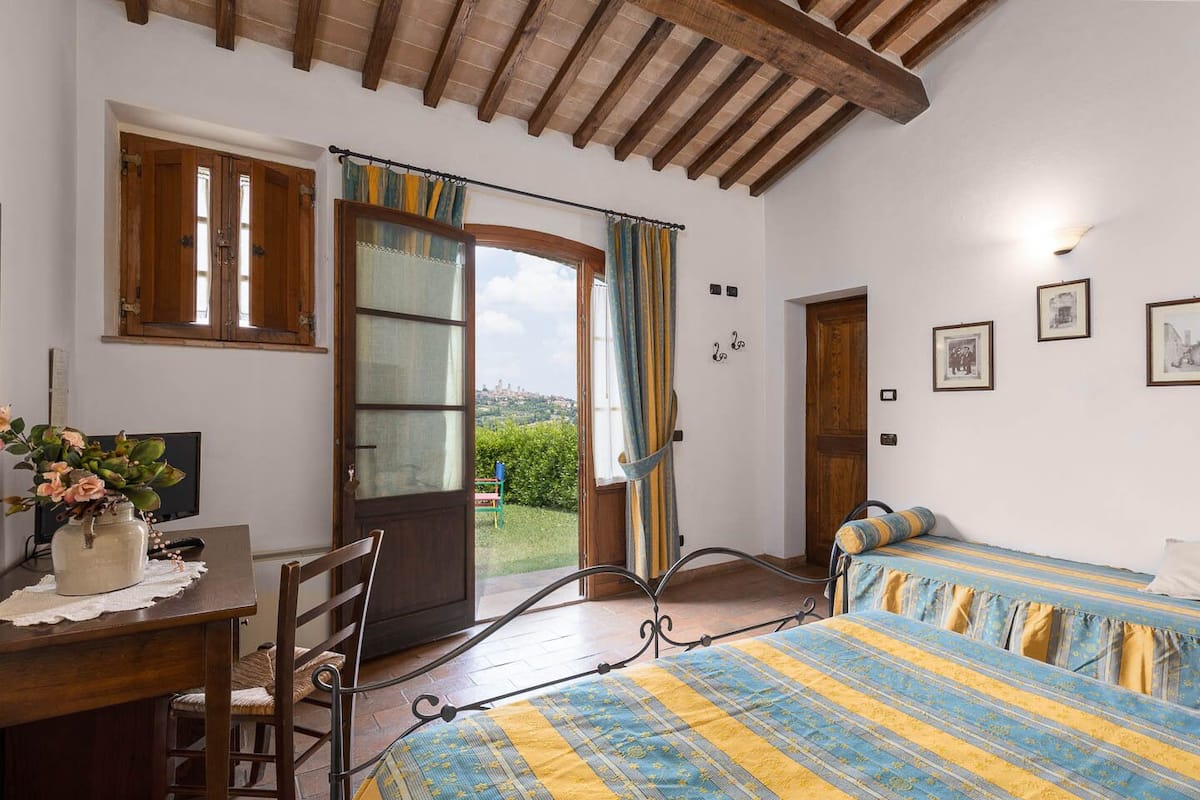 Quarto com cama, escrivaninha, cadeira e janela com cortinas.