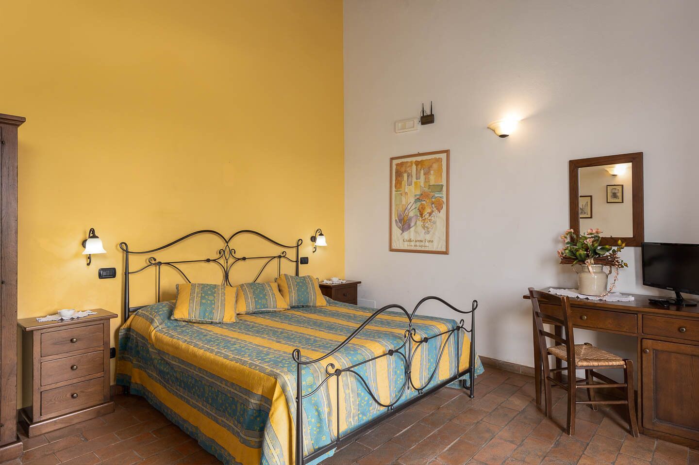A bedroom with a bed, bedside tables, a desk with a chair, a television, and a wall-mounted light fixture.