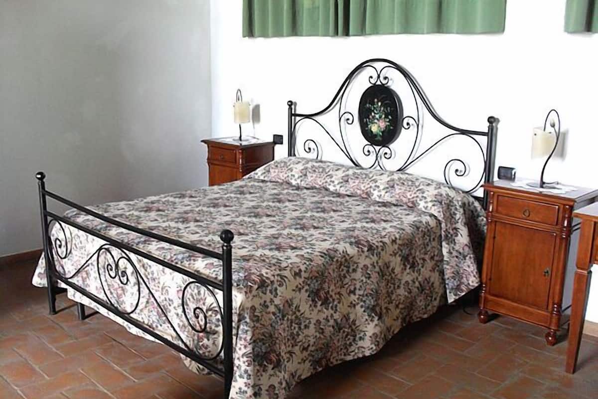 Uma cama com colcha florida, mesa de cabeceira de madeira e luminária fixada na parede.