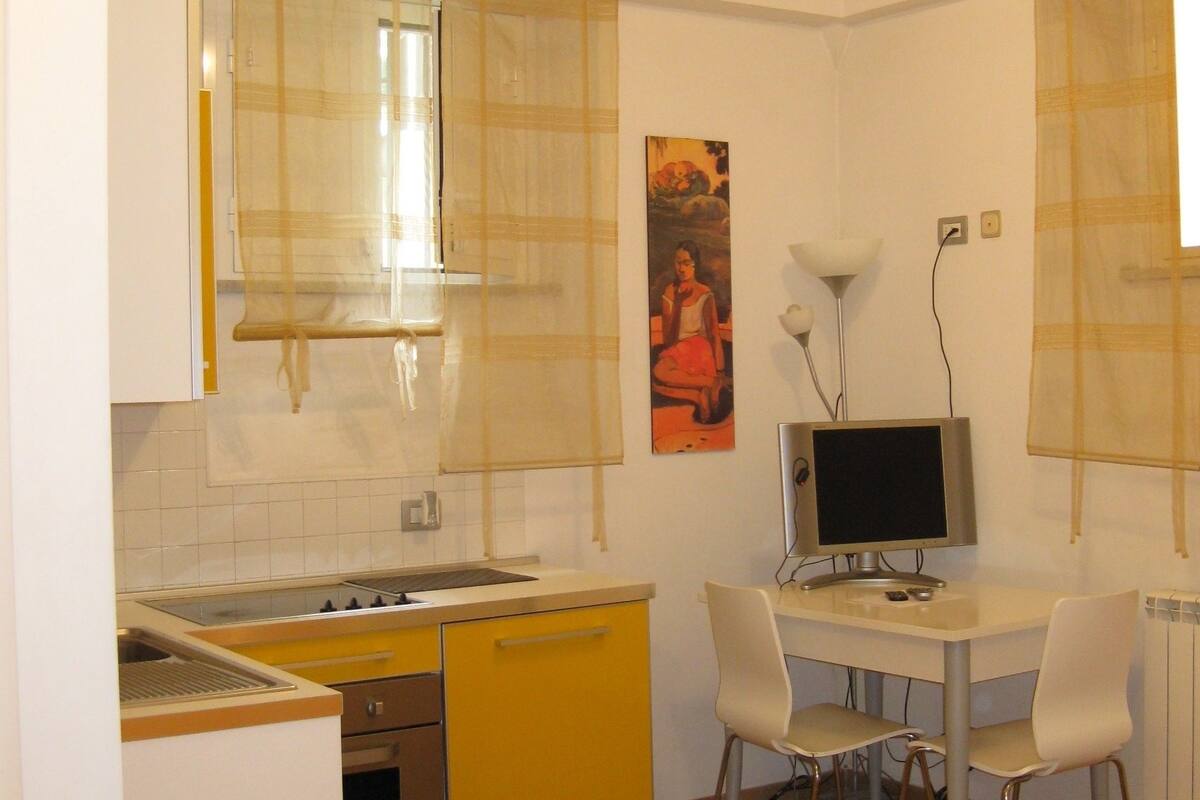 A compact kitchen with a stove, sink, and yellow cabinets. There is a dining area with a table and two chairs, and a bed with two folded towels.