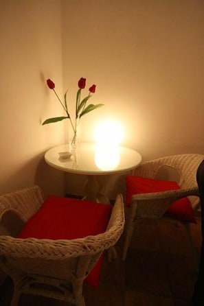 Ein kleiner runder Tisch mit einer Rosenvase, zwei Korbstühle mit roten Kissen und eine beleuchtete Lampe.