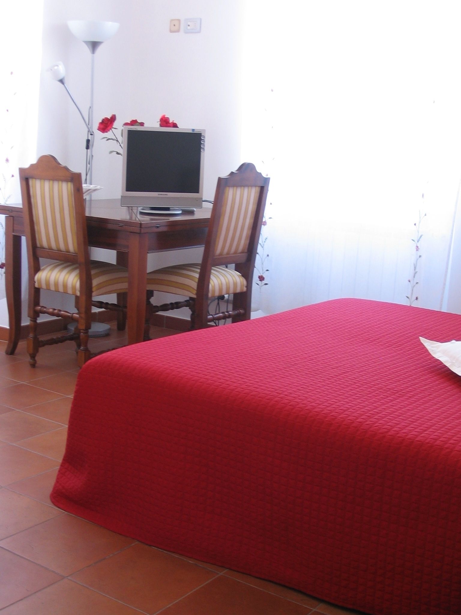 Una habitación con cama, dos sillas, un escritorio con computadora y una lámpara.