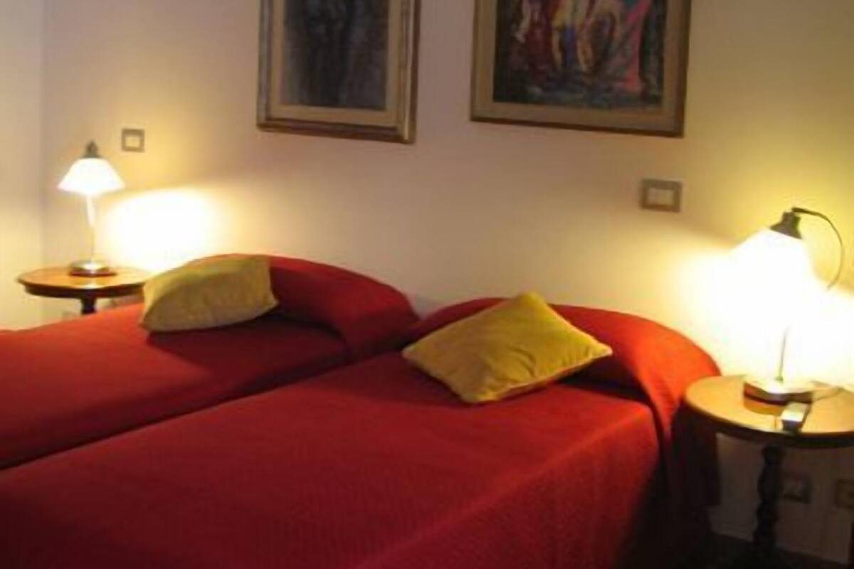 A room with two beds, each with a red bedspread and a yellow pillow. There are two lamps on side tables, and two framed artworks on the wall.