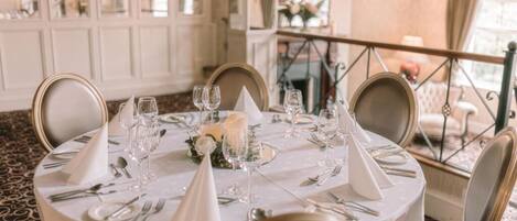 A round dining table set with white tablecloth, silverware, and glassware, with a floral centerpiece.