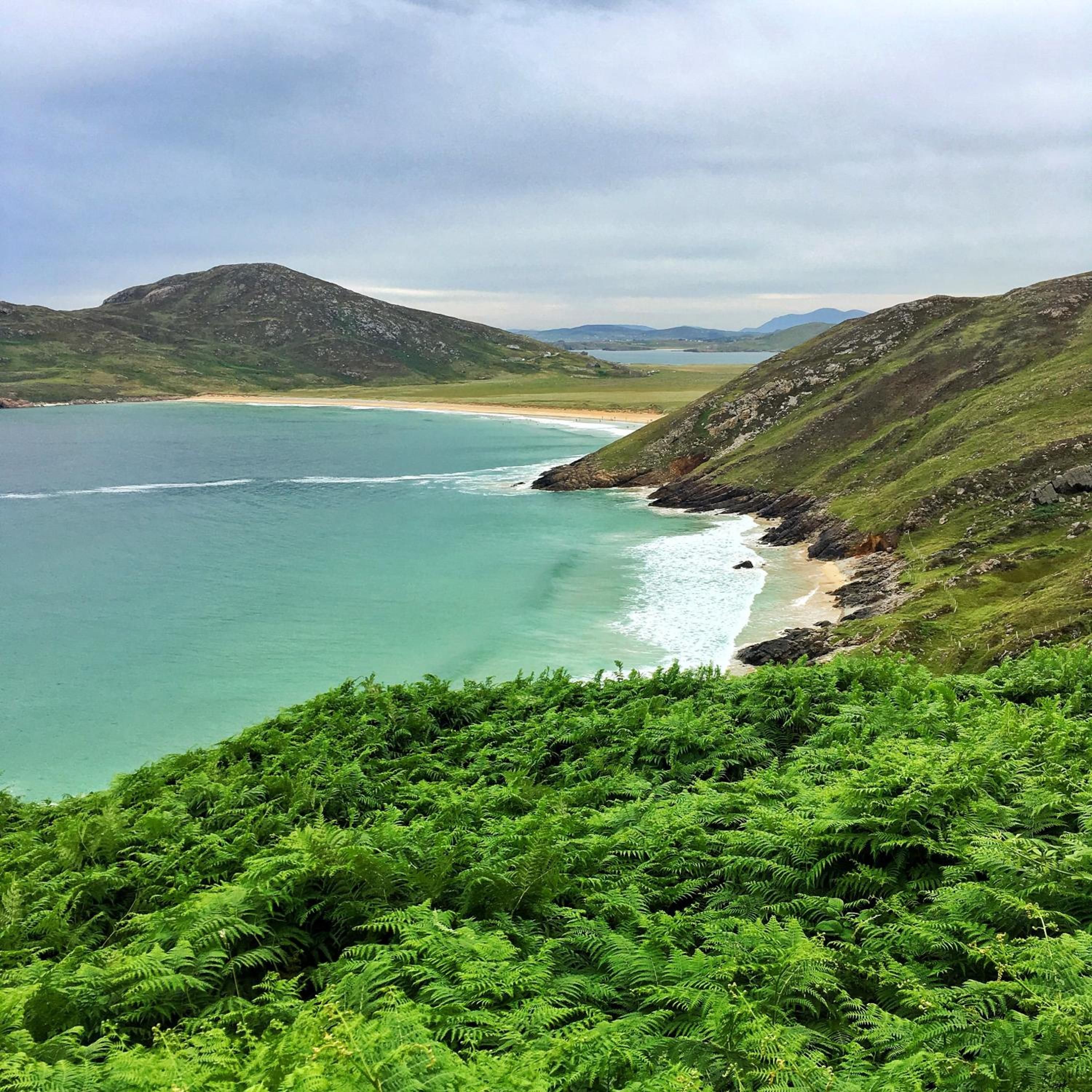A coastal landscape with a beach, green hills, and a clear sky.