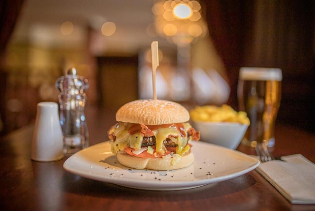 Una hamburguesa en un plato con un tenedor y cuchillo a un lado, una jarra de cerveza y un bol de papas fritas al fondo.