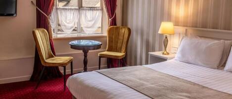 A hotel room with a bed, two chairs, a small table, a lamp, and a television on the wall.