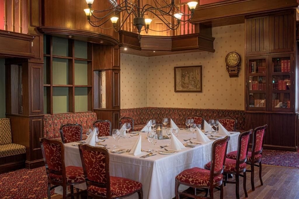 Comedor formal con techo alto, paneles de madera ornamentados y una araña. Las mesas están dispuestas con manteles blancos y sillas tapizadas en rojo.