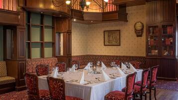 Comedor formal con techo alto, paneles de madera ornamentados y una araña. Las mesas están dispuestas con manteles blancos y sillas tapizadas en rojo.