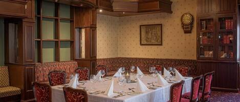 Comedor formal con techo alto, paneles de madera ornamentados y una araña. Las mesas están dispuestas con manteles blancos y sillas tapizadas en rojo.