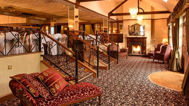 A grand room with a patterned carpet, a wooden staircase, and a fireplace. - Kee's Hotel  (Stranorlar)