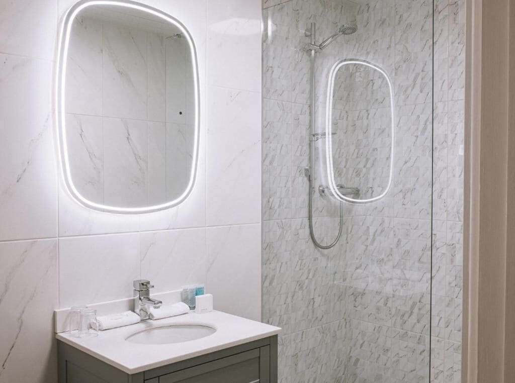 A bathroom with a marble-tiled wall, a sink with a mirror, and a shower with a handheld showerhead.