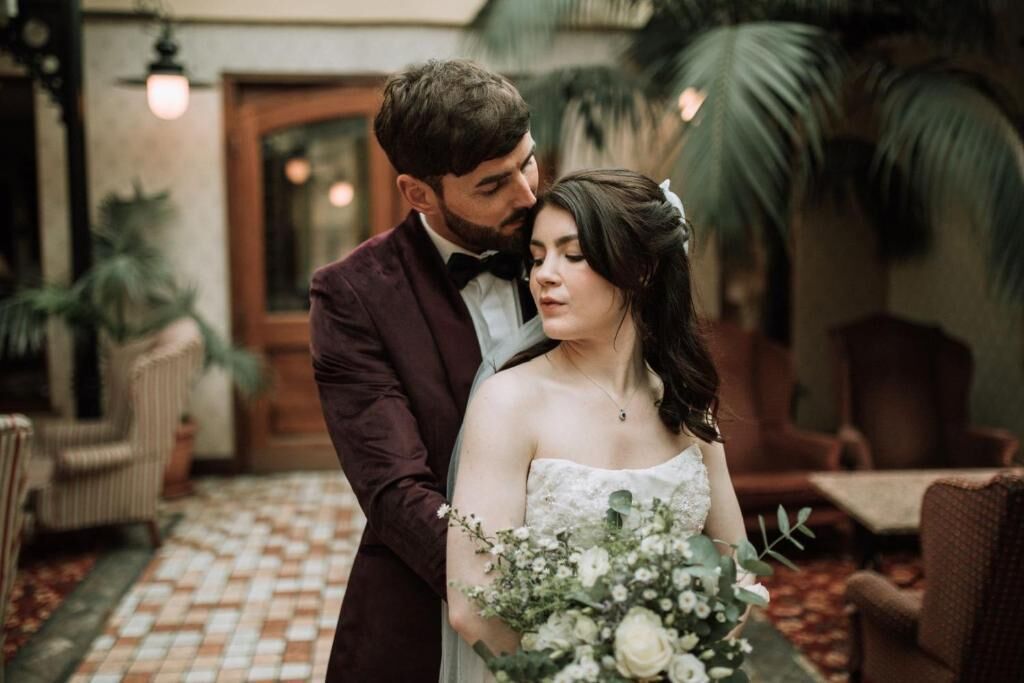 A couple in a formal setting with a patterned floor, wicker furniture, and indoor plants.