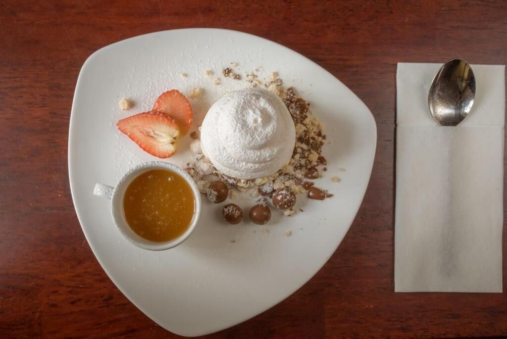 Un plato blanco con un postre que incluye una porción de crema batida, salsa de caramelo y nueces y trozos de chocolate esparcidos.