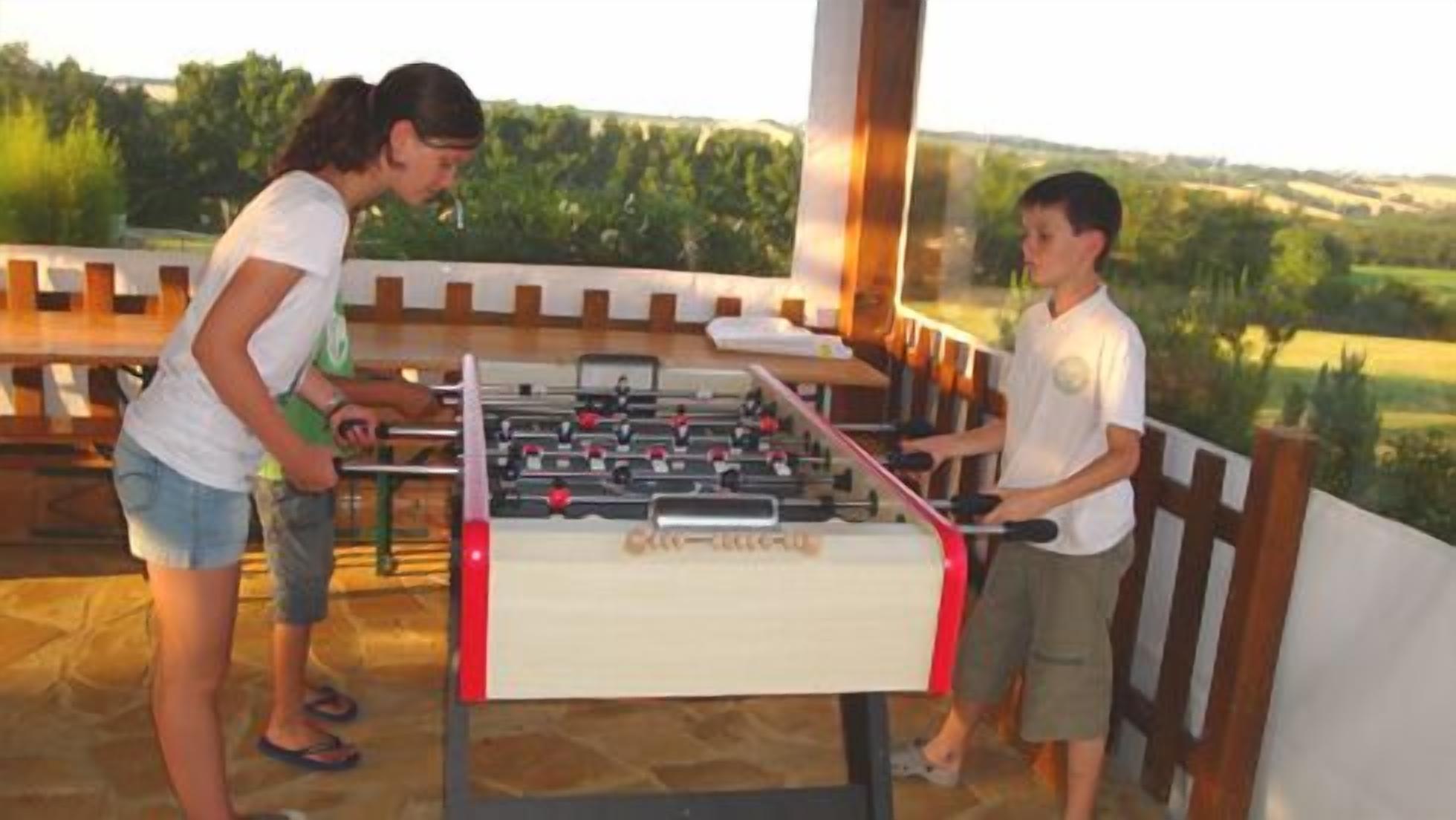 Een tafelvoetbaltafel met twee spelers, een volwassene en een kind, buiten met uitzicht op groen en heuvels.