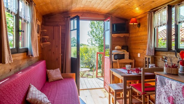 L’intérieur d’un chalet accueillant, avec un plafond en bois, un canapé rouge agrémenté de coussins à motifs, une table à manger avec des chaises, et une vue sur la verdure à l’extérieur.