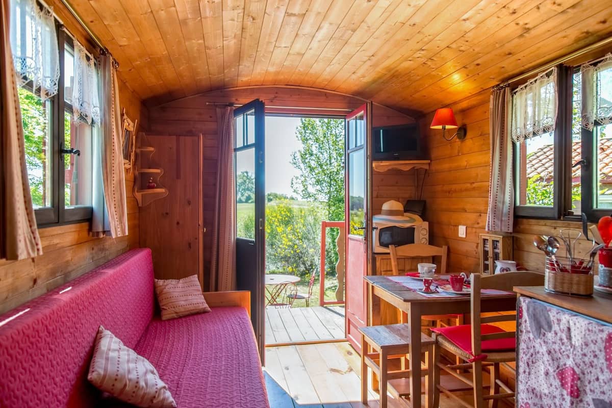 Een knus interieur van een blokhut, met een houten plafond, een rode bank met opvallende kussens, een eettafel met stoelen en uitzicht op groen buiten.