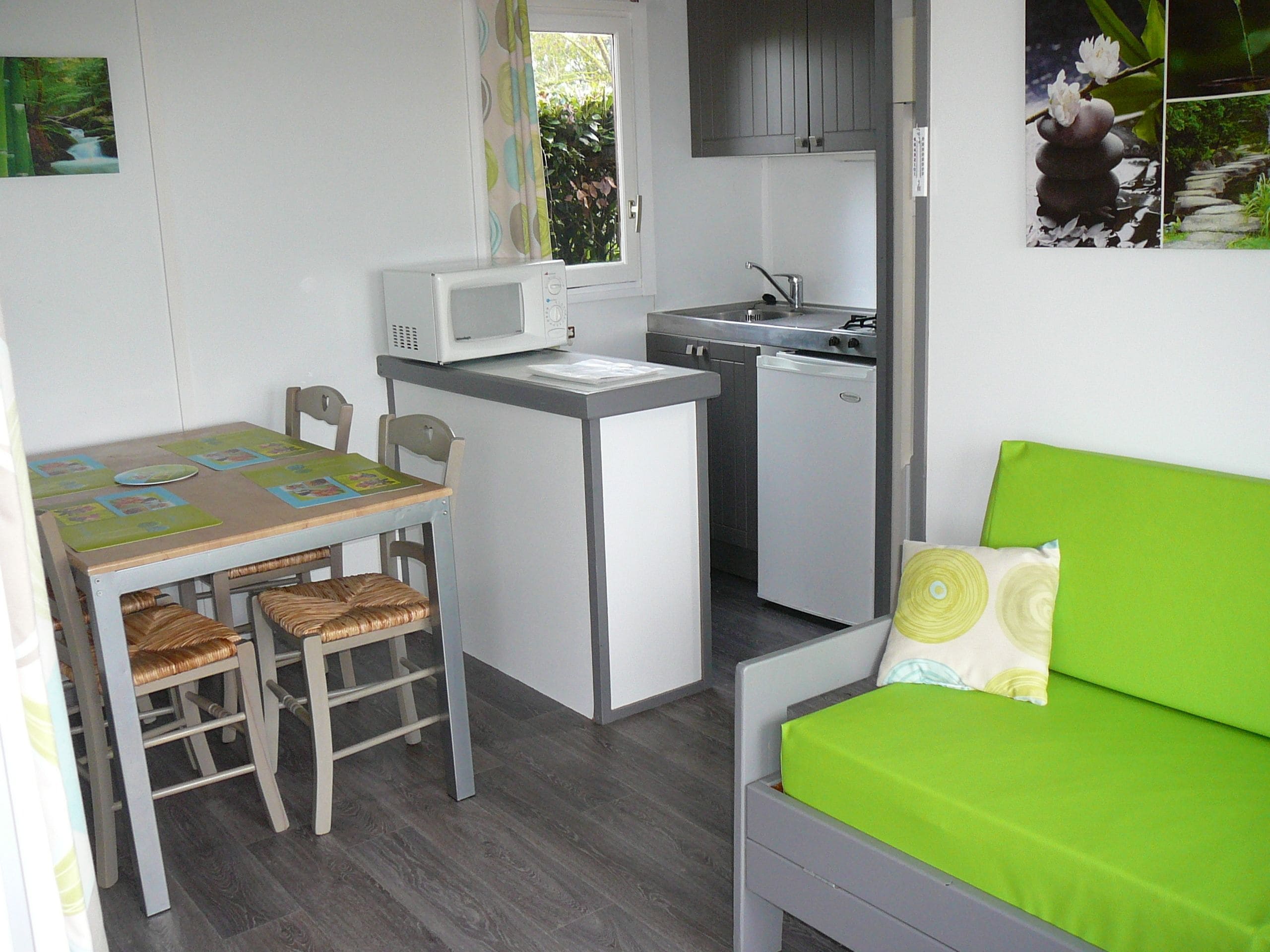 A compact kitchen with a microwave, sink, and small refrigerator. There is a dining table with chairs and a green sofa with a pillow.