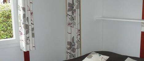 A single bed with a grey bedspread and two pillows, a window with white curtains, and a white wardrobe with a door partially open.