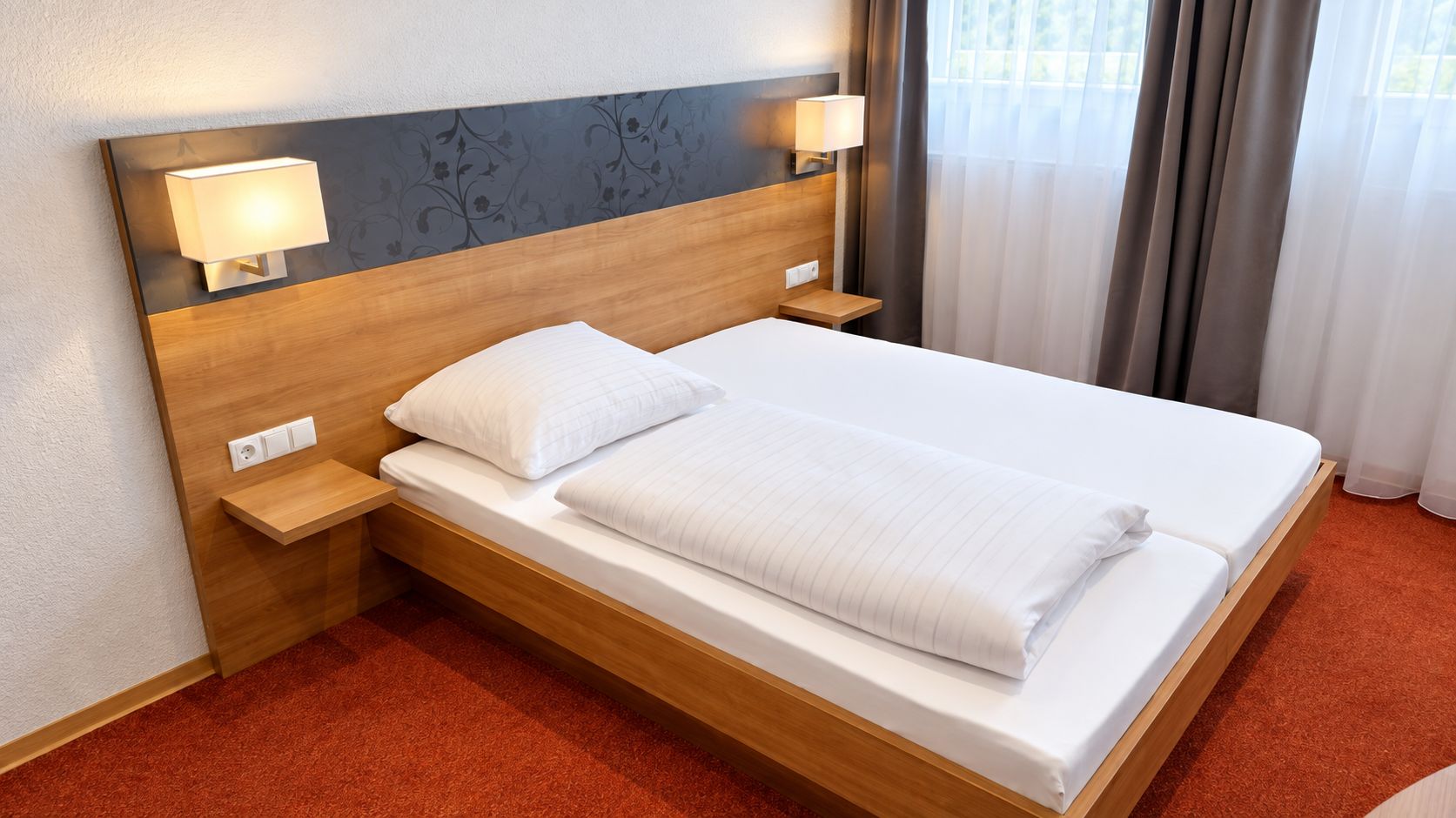 Habitación de hotel con cabecera de madera, una cama individual con ropa blanca, dos lámparas de noche y una alfombra roja.