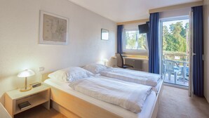 A hotel room with two beds, a desk, a chair, and a balcony with a view of trees. - HRC Pfeffermühle (Landstuhl)