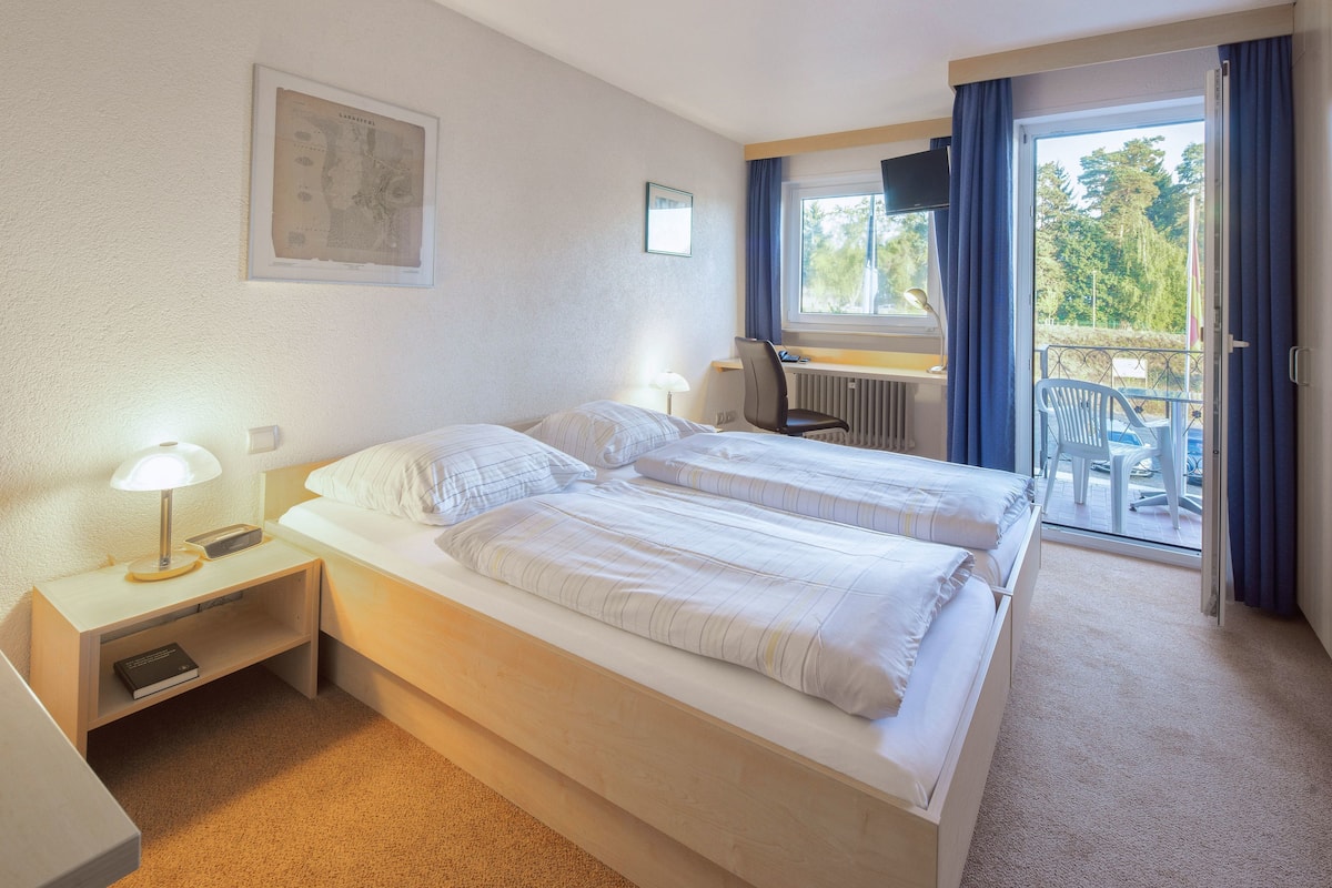 A hotel room with two beds, a desk, a chair, and a balcony with a view of trees.