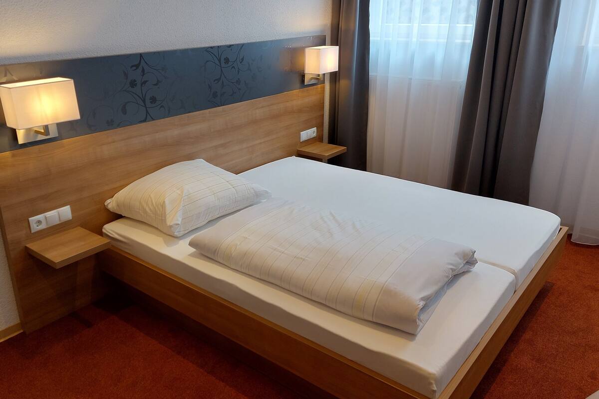 A hotel room with a wooden headboard, two bedside lamps, a neatly made bed with white linens, and a carpeted floor.