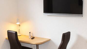 Una habitación pequeña con televisión de pantalla plana montada en la pared, una mesa con dos sillas, una lámpara y un equipo de aire acondicionado.