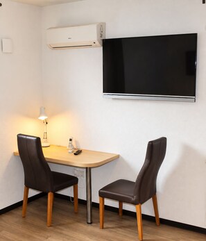 Una habitación pequeña con televisión de pantalla plana montada en la pared, una mesa con dos sillas, una lámpara y un equipo de aire acondicionado.