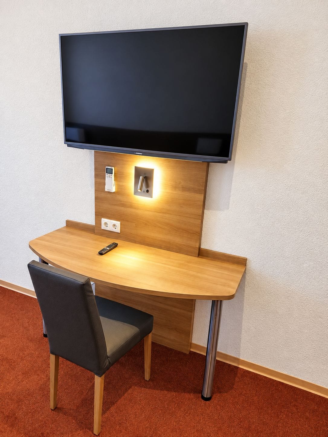 A modern desk with a flat-screen TV mounted above it, a remote control on the desk, and a chair with a cushioned seat.