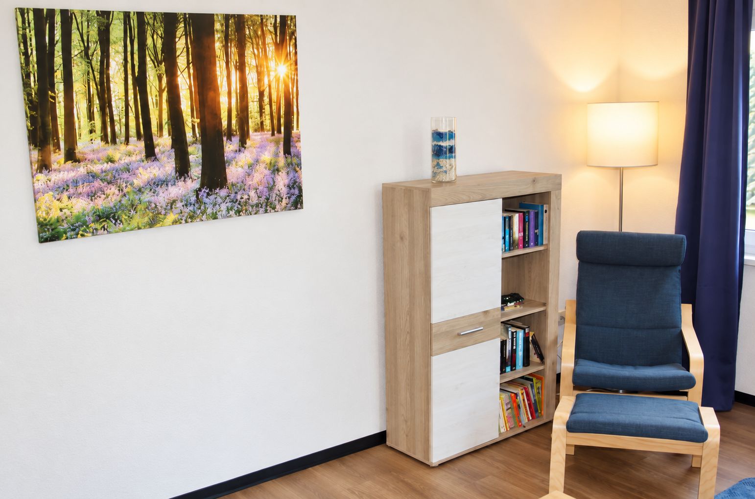 A room with a bookshelf, a chair, a lamp, and a painting of a forest.
