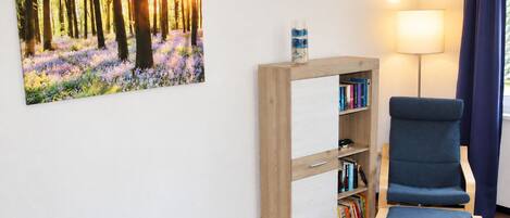 Una habitación con una estantería, una silla, una lámpara y un cuadro de un bosque.