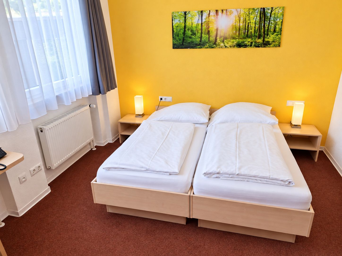 A hotel room with a double bed, two bedside tables with lamps, a radiator, a window with curtains, and a forest-themed wall art.