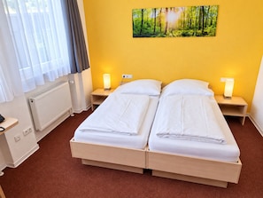 Una habitación de hotel con una cama doble, dos mesitas de noche con lámparas, un radiador, una ventana con cortinas y una obra de arte en la pared con temática de bosque.