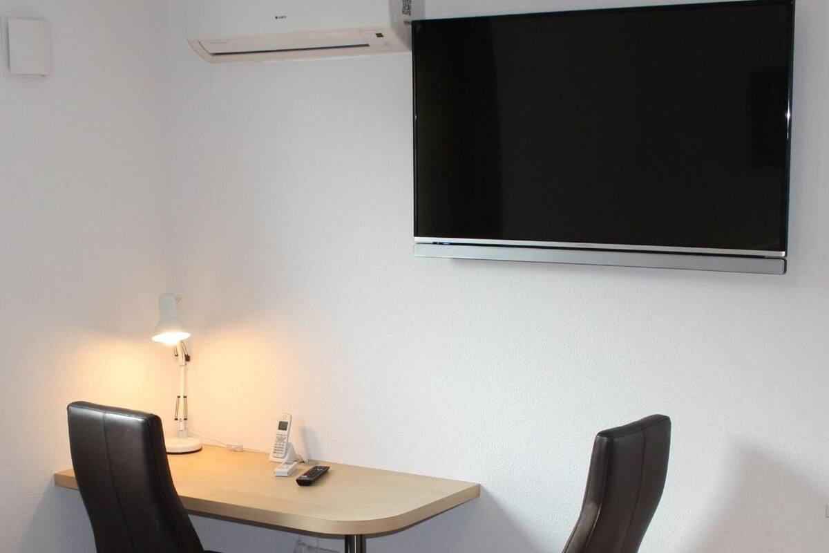A room with a TV mounted on the wall, a table with two chairs, a lamp, and a phone.