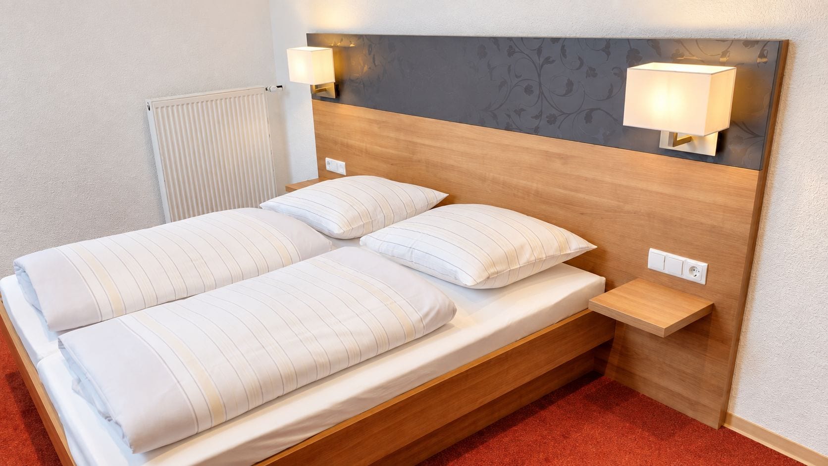 Una cama doble con dos almohadas, una mesita de noche, una lámpara de pared y un radiador.