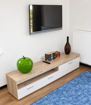 Una sala de estar moderna con un televisor de pantalla plana montado en la pared, una mesa consola de madera con una manzana verde, libros y un jarrón, además de una alfombra azul estampada.